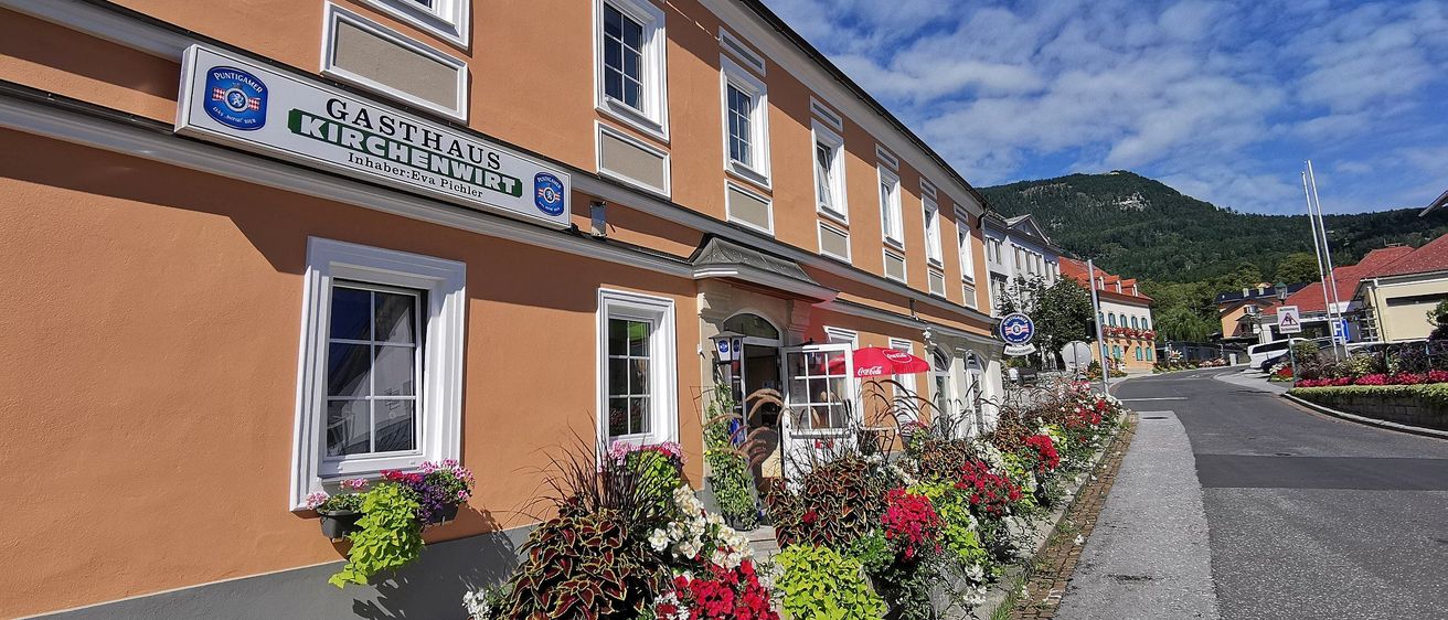 Eine Reihe von Gebäuden hat ein Schild mit der Aufschrift Gasthaus Kirchenwirt. Es hat viele Fenster und Topfpflanzen. Davor stehen Blumen.