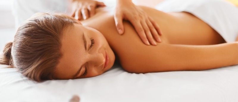 Eine Frau liegt auf dem Bauch auf einem weißen Bett und erhält eine Rückenmassage von einer Masseurin.