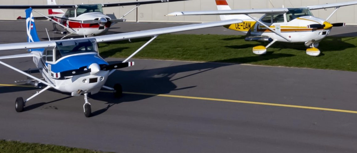 Zwei kleine Flugzeuge sind auf dem Rollfeld eines Flughafens geparkt. Ein Flugzeug hat das Kennzeichen UE-DAW und ein anderes Flugzeug hat einen roten Heckausleger.
