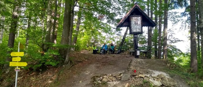 Zwei Personen sitzen auf Bänken in einem Waldgebiet. Eine kleine Kapelle mit einem Glasfenster befindet sich in der Nähe.