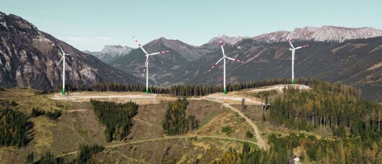 Luftaufnahme von vier Windturbinen, die auf einem Bergkamm installiert sind, mit einem bewaldeten Gebiet darunter und einer Bergkette im Hintergrund.