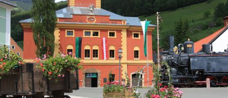 Ein leuchtend rotes Gebäude mit gelben Akzenten, einer Statue oben und Flaggen steht in der Nähe eines Bahnhofs. Ein schwarzer Zug ist vor dem Gebäude geparkt. Bunte Blumen schmücken die Umgebung.