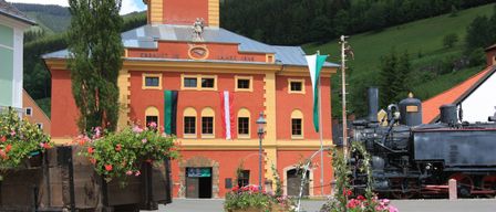 Ein leuchtend rotes Gebäude mit grauem Dach, das mit Flaggen und einer Statue geschmückt ist, steht neben einer Dampflokomotive und ist von blühenden Pflanzen umgeben.