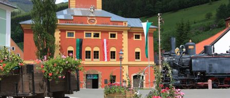 Ein großes rotes Gebäude mit gelben Akzenten, das eine Statue und Flaggen aufweist, steht vor einer bergigen Kulisse. Davor schmücken eine alte Lokomotive und ein blumenreicher Wagen die Szene.