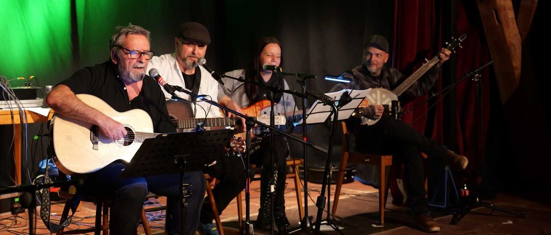 Vier Musiker stehen auf der Bühne und spielen vor einem grünen Vorhang. Sie spielen Gitarren und singen in Mikrofone. Die Bühne ist schwach beleuchtet.