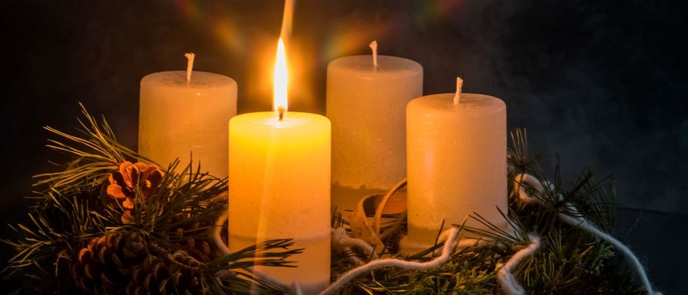 Vier brennende Kerzen umgeben von Kiefernzapfen und einer Tannenzapfen. Die Kerzen stehen vor einem dunklen Hintergrund mit einem Lichteffekt.
