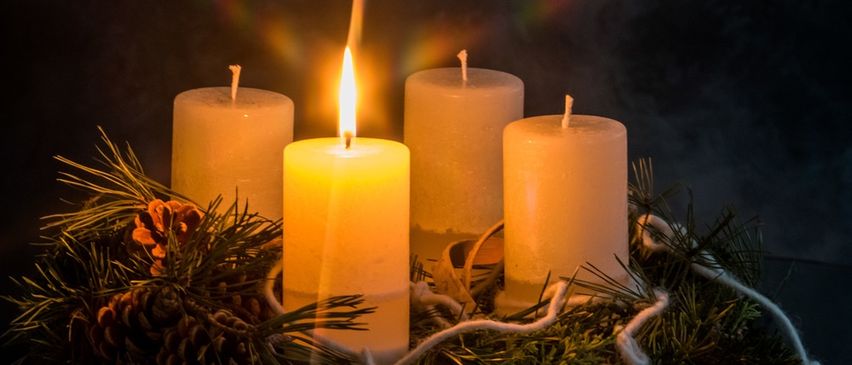 Vier brennende Kerzen umgeben von Kiefernzapfen und einer Tannenzapfen. Die Kerzen stehen vor einem dunklen Hintergrund mit einem Lichteffekt.