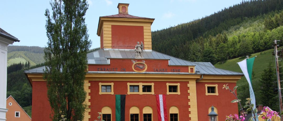 Ein rotes Gebäude mit gelbem Rand hat Statuen auf dem Dach und Flaggen an den Fenstern. Das Gebäude ist von Blumen umgeben.