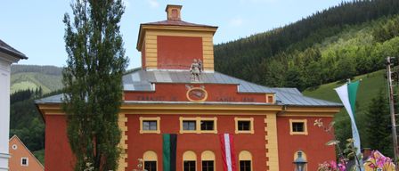 Ein rotes Gebäude mit gelbem Rand hat Statuen auf dem Dach und Flaggen an den Fenstern. Das Gebäude ist von Blumen umgeben.