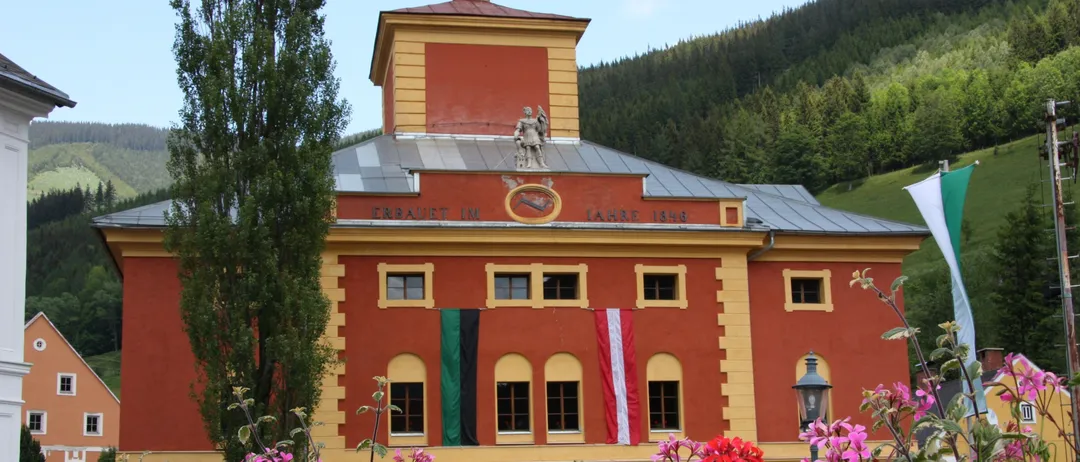 Ein großes rotes Gebäude mit gelben Akzenten steht zwischen Bäumen und Blumen. Es hat einen Turm mit einer Statue oben, und Fahnen hängen an den Fenstern. Das Schild des Gebäudes lautet 'ERBAUT IM JAHRE 1848'.