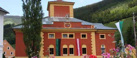 Ein großes rotes Gebäude mit gelben Akzenten steht zwischen Bäumen und Blumen. Es hat einen Turm mit einer Statue oben, und Fahnen hängen an den Fenstern. Das Schild des Gebäudes lautet 'ERBAUT IM JAHRE 1848'.