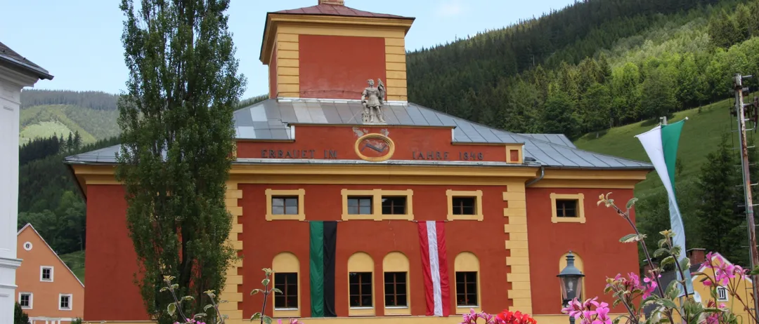 Ein großes rotes Gebäude mit gelben Akzenten und einem Turm, das Flaggen und ein Schild mit der Aufschrift 'Erbaut im Jahre 1846' zeigt. Umgeben von leuchtenden Blumen und einem hohen Baum. Im Hintergrund befinden sich üppige grüne Hügel.