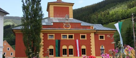 A large red building with yellow accents and a tower, displaying flags and a sign reading 'Erbaut im Jahre 1846.' Surrounded by vibrant flowers and a tall tree. In the background, there are lush green hills.