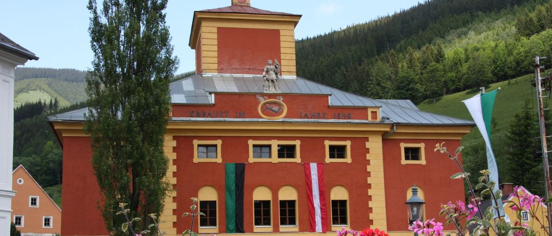 Ein rotes Gebäude mit gelbem Rand, das Flaggen zeigt und von Blumen umgeben ist. Eine Statue oben und der Text 'ERBAUET IM JAHRE 1846'.