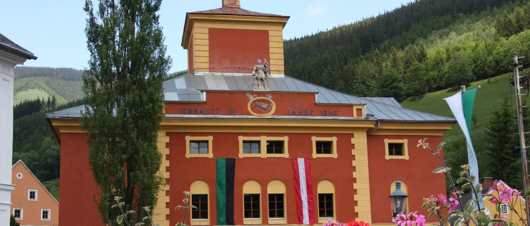 Ein rotes Gebäude mit gelben Akzenten und einem Turm steht vor einem Hügel. Es hat mehrere Fenster und Flaggen und Blumen davor.