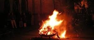 Ein Lagerfeuer brennt hell in der Nacht, mit Leuten, die darum stehen, und einem Gebäude mit Bogenfenstern im Hintergrund.