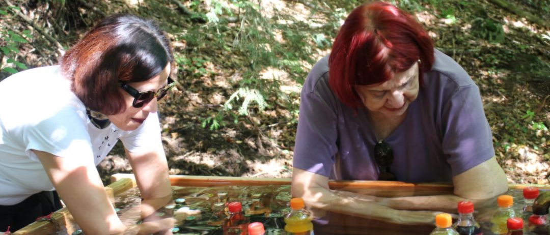 Zwei Frauen untersuchen eine Holzkiste mit Wasser, die verschiedene Flaschen mit orangefarbenen Kappen enthält. Die Szene spielt in einem Waldgebiet.