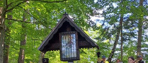 Eine Gruppe von Menschen steht in einem Wald, zwei von ihnen in Uniformen, und einer salutiert. Hinter ihnen befindet sich ein Gedenkschrein.