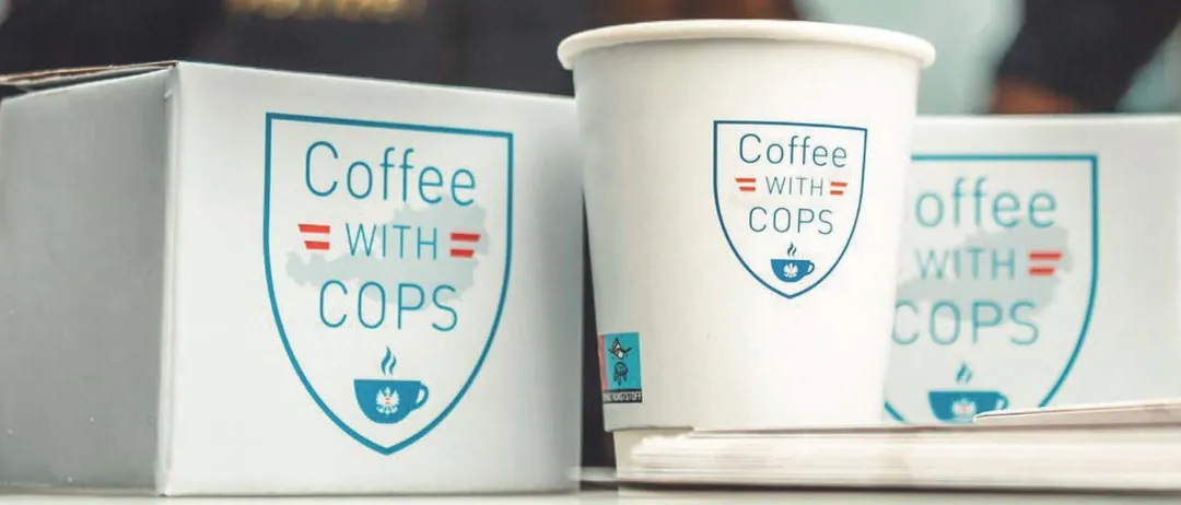 Zwei Polizisten lächeln, einer hält eine Kaffeetasse, unter einem Zelt mit 'Kaffee mit Cops' Branding.