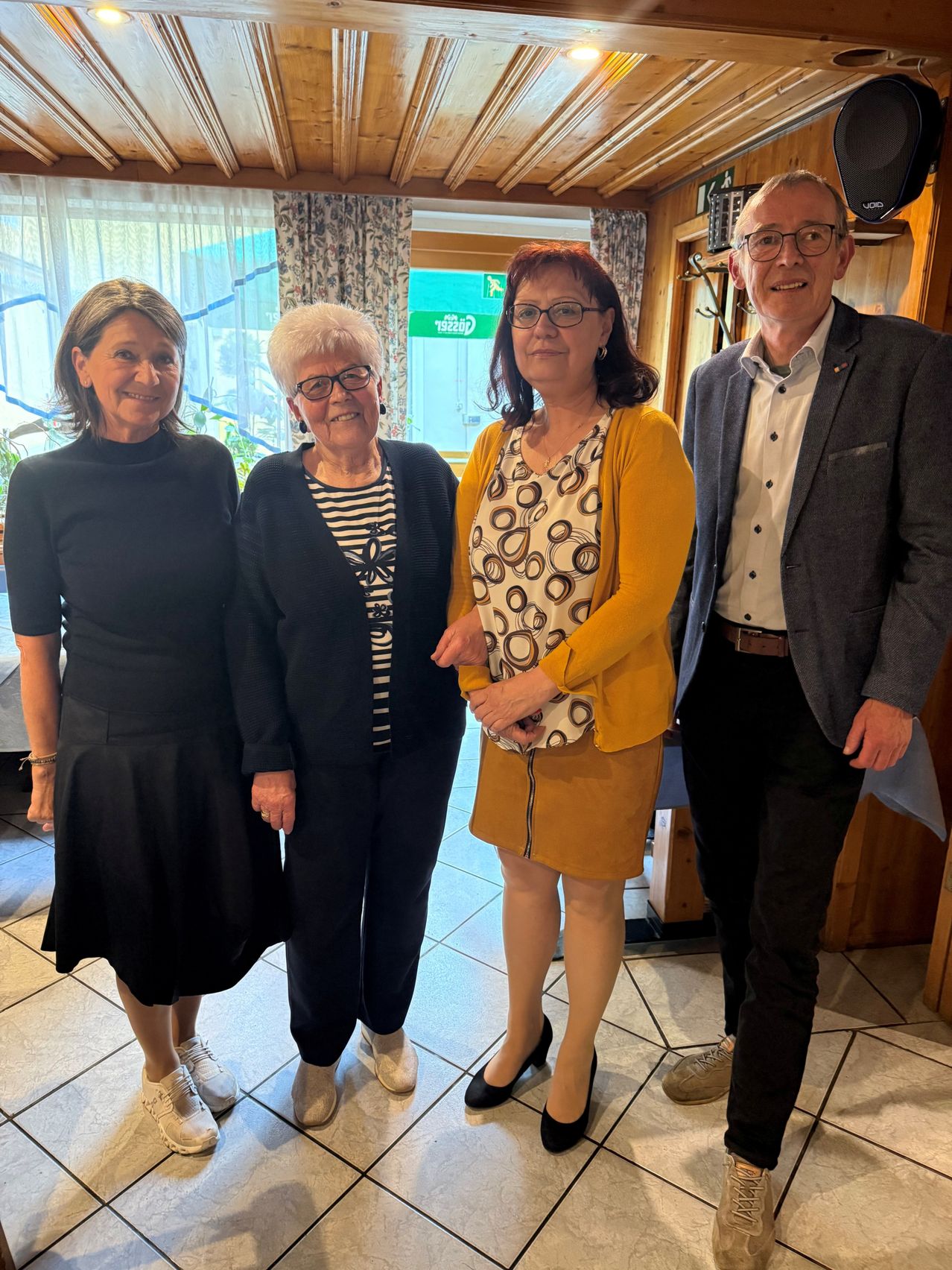 Vier Erwachsene, zwei Frauen und zwei Männer, stehen eng zusammen und lächeln für ein Foto. Sie sind lässig gekleidet, wobei zwei Frauen eine Brille tragen. Hinter ihnen ist eine Holzwand und ein Fenster mit Vorhängen zu sehen.