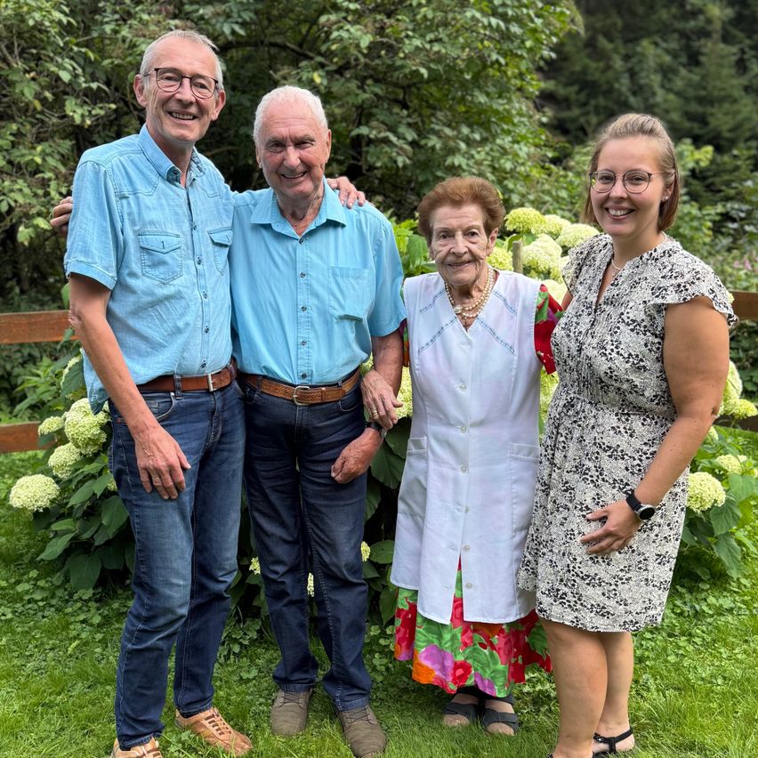 Vier Personen stehen in einem Garten mit weißen Blumen und grünen Pflanzen. Zwei ältere Männer und zwei Frauen, eine ältere und eine junge. Sie alle lächeln in die Kamera.