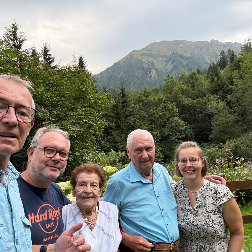 Vier Menschen posieren für ein Foto vor einer Berglandschaft. Die beiden älteren Männer und Frauen lächeln, und der jüngere Mann und die Frau haben ihre Arme um sie herum.