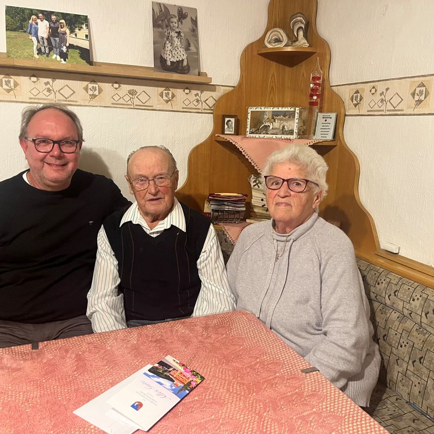 Drei Menschen sitzen lächelnd an einem Tisch. Ein Mann und eine Frau sitzen auf beiden Seiten eines älteren Mannes. Eine rosa Tischdecke und einige Karten liegen auf dem Tisch.