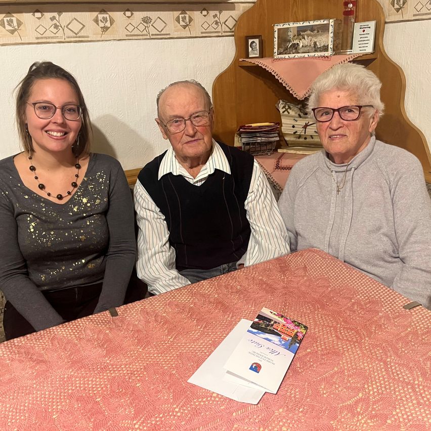 Drei Personen sitzen um einen Tisch herum. Eine lächelnde Frau links trägt eine Brille und eine Halskette. Ein älterer Mann in der Mitte trägt eine Brille und eine Weste. Eine ältere Frau rechts trägt eine Brille und eine graue Sweatshirt.