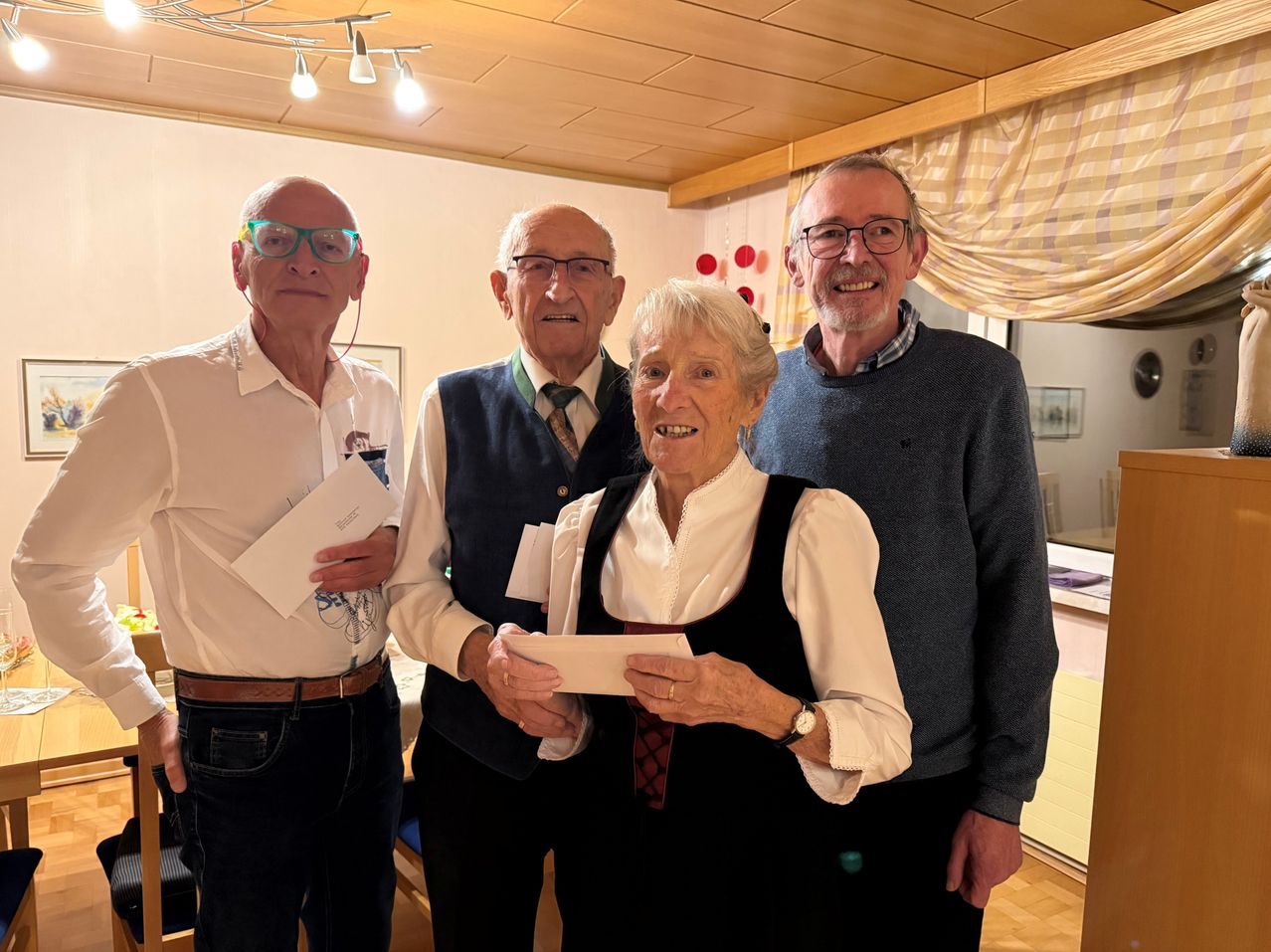 Vier Menschen stehen lächelnd in einem Raum. Eine ältere Frau hält ein Stück Papier, während ein älterer Mann neben ihr ein weiteres hält. Zwei Männer stehen auf beiden Seiten, einer hält ein Stück Papier und der andere trägt eine Brille.