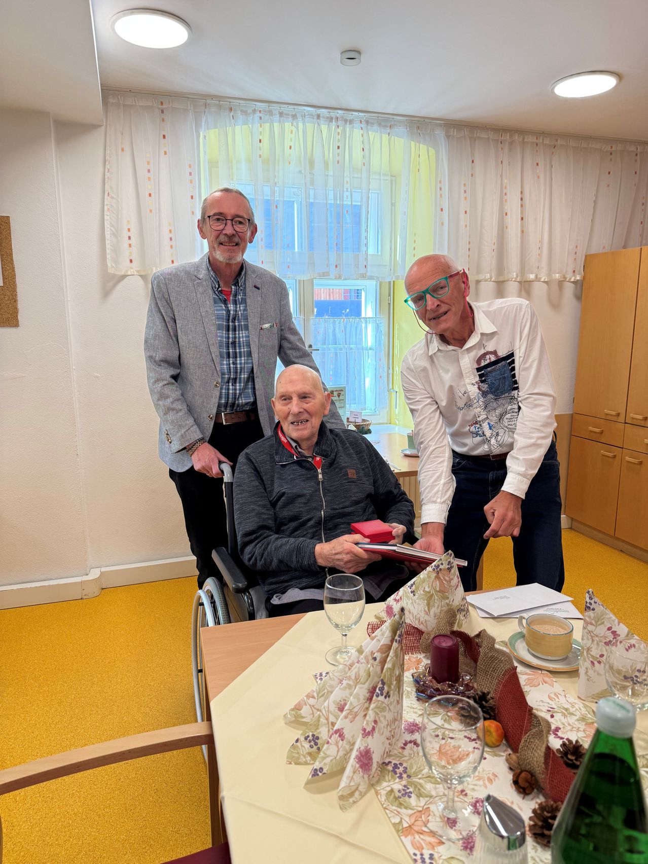 Drei Männer stehen um einen Tisch herum, einer im Rollstuhl. Der Mann im Rollstuhl hält ein Buch, und die anderen lächeln. Der Raum hat ein Fenster mit Vorhängen und einen gelben Boden.