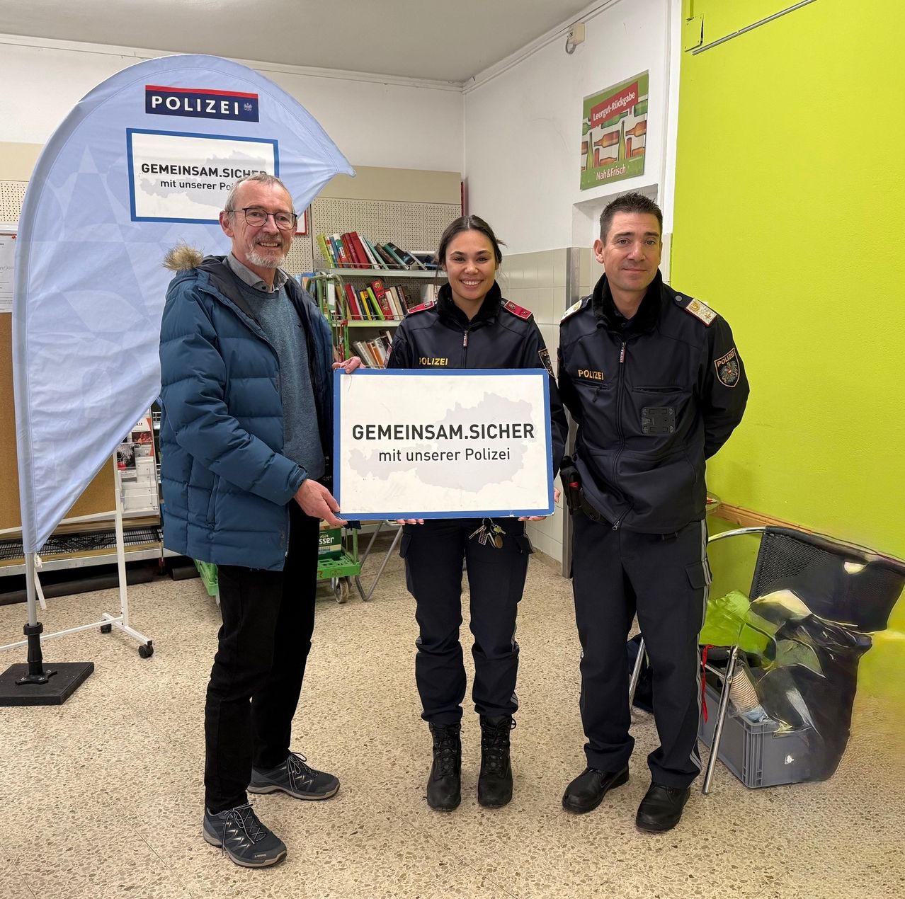Drei Personen stehen lächelnd in einem Gebäude. Ein Mann in einer blauen Jacke hält ein Schild mit der Aufschrift 'GEMEINSAM.SICHER mit unserer Polizei.' Die Frau neben ihm trägt eine Polizeiuniform, ebenso wie der Mann rechts. Ein Bücherregal und ein blauer Banner mit dem Wort 'POLIZEI' sind hinter ihnen.