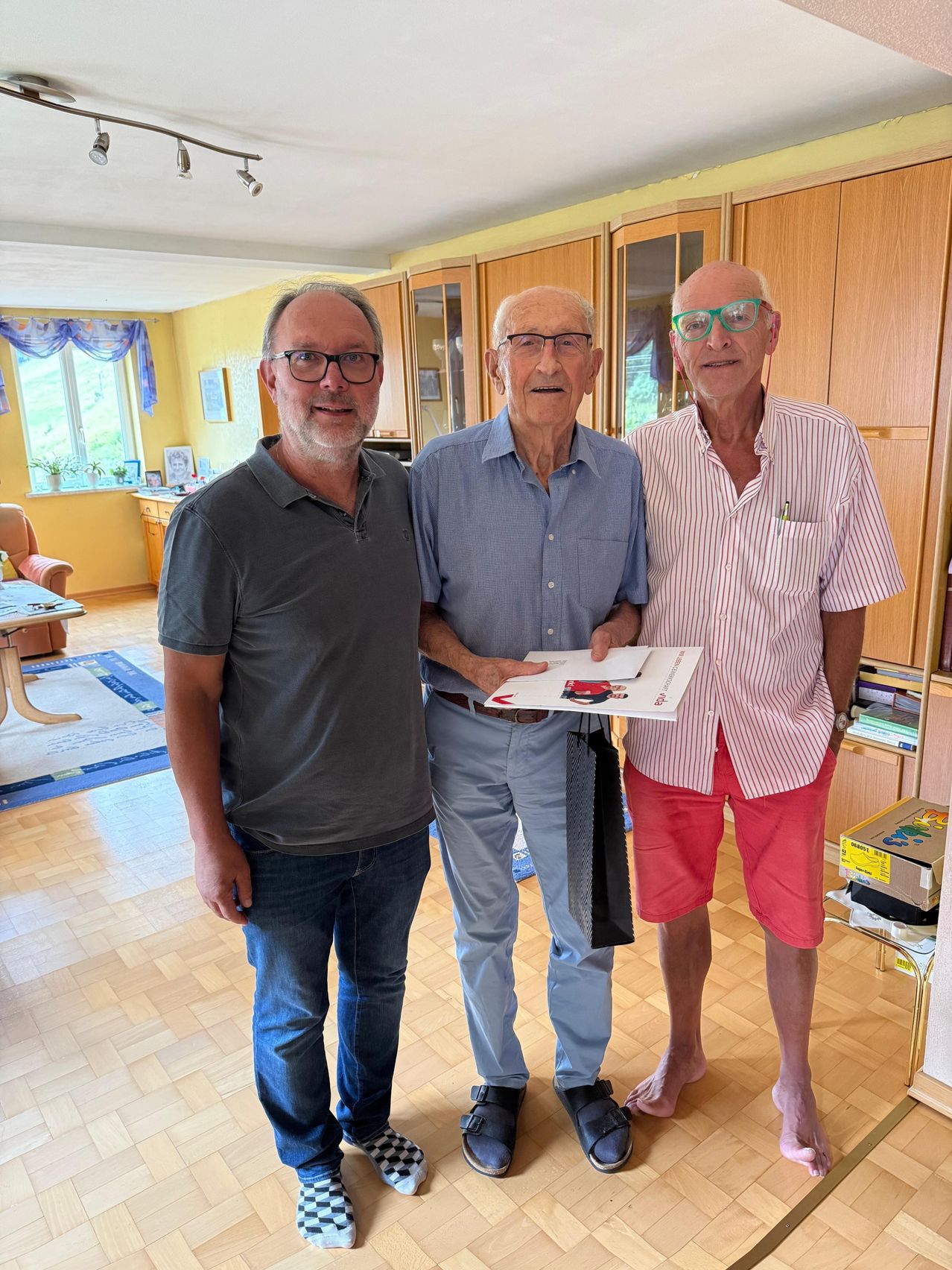 Three men stand in a room, the man in the middle holds a book, while the man on the right wears glasses and a striped shirt.