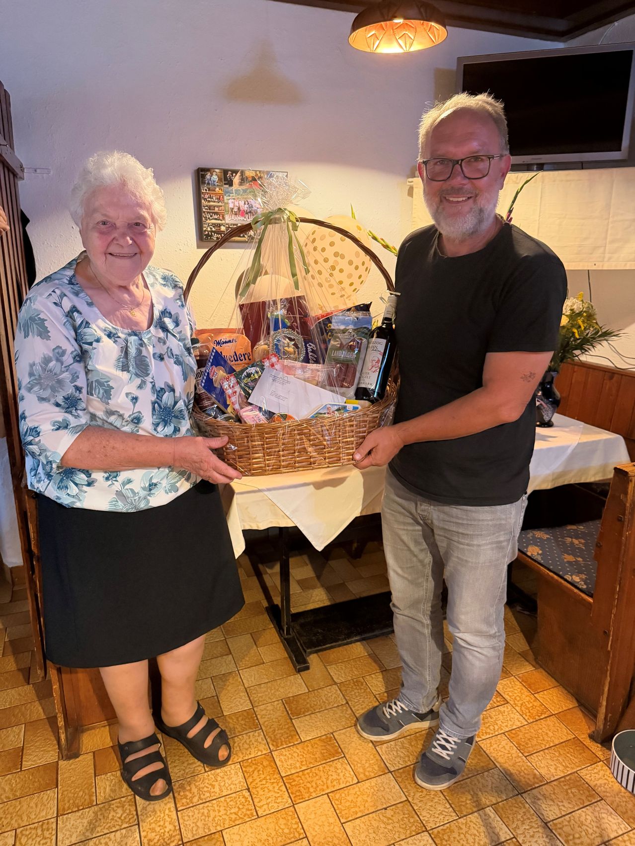 An elderly woman and a man are holding a basket filled with various items, possibly a gift basket, in a room.