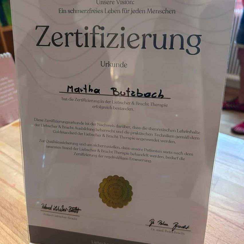 A certificate on a wooden table, titled 'Zertifizierung' in German, with the name 'Martha Butzbach' and a gold seal. It states that Martha has successfully completed the certification in Liebscher & Bracht Therapy. The certificate highlights the importance of theoretical knowledge and practical techniques in the therapy.
