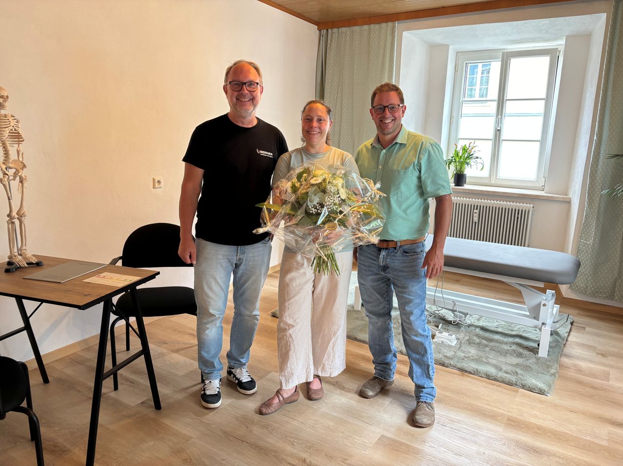 Three people stand in a room with a bouquet of flowers. The woman in the middle holds the bouquet. The man on the left wears glasses and a black shirt. The man on the right wears a green polo and jeans. Behind them is a window with a potted plant.