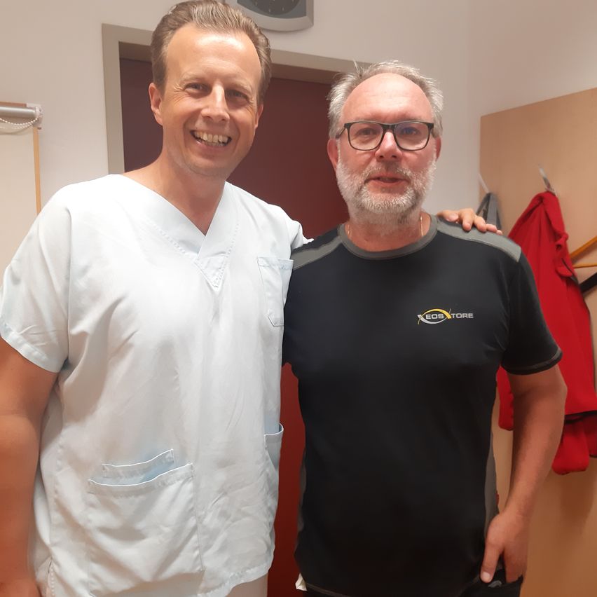 Zwei Männer stehen zusammen, einer in einem weißen Laborkittel und der andere in einem schwarzen T-Shirt mit Logo. Sie lächeln und posieren für ein Foto in einem Raum.