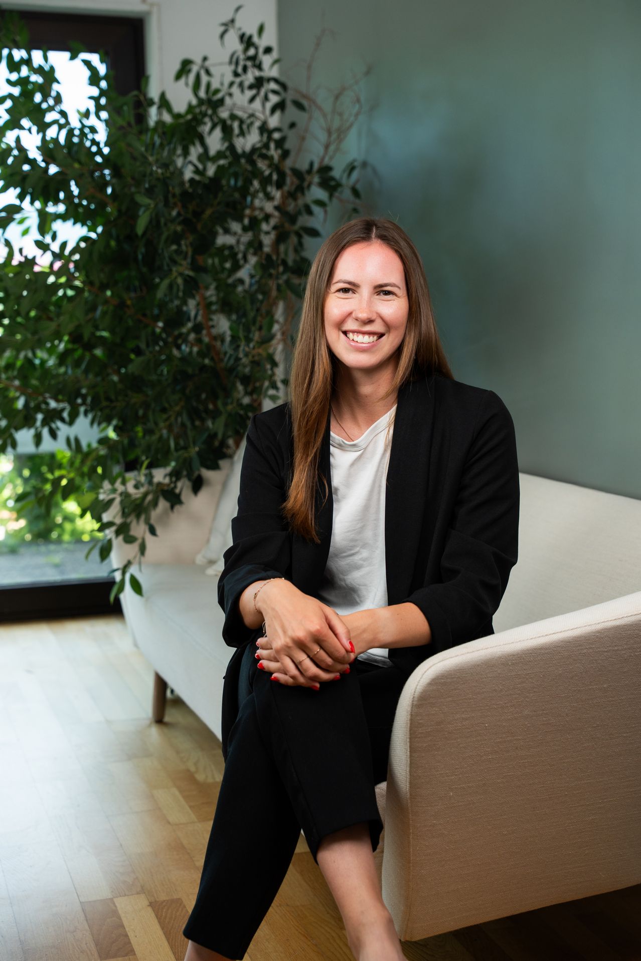 A smiling woman in a black blazer and white shirt sits on a white couch, her hands clasped together, with a large plant in the background.