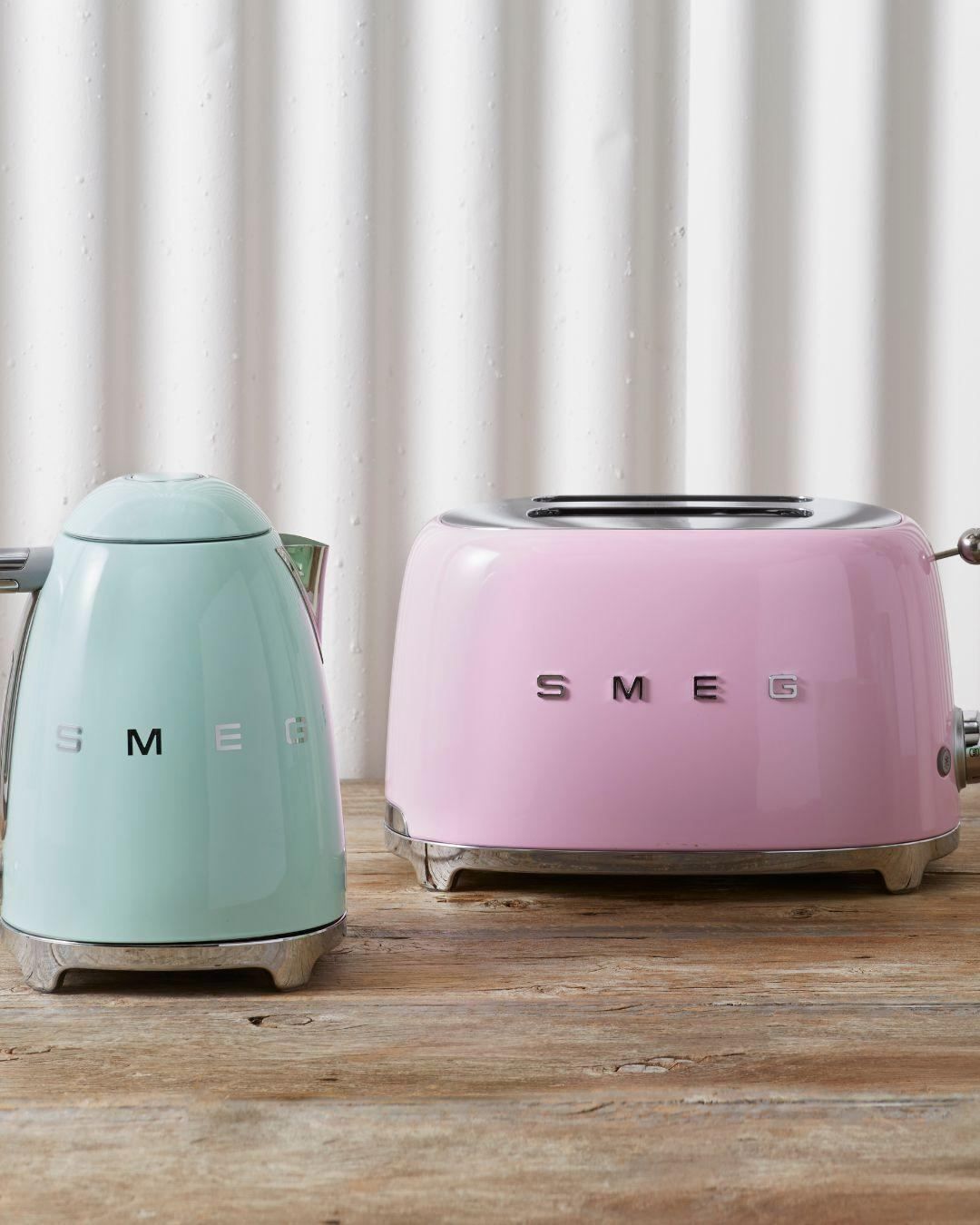 Two pastel-colored Smeg kitchen appliances, a kettle and a toaster, are displayed on a wooden surface. The kettle is mint green, and the toaster is pink. Both have the Smeg logo on them.