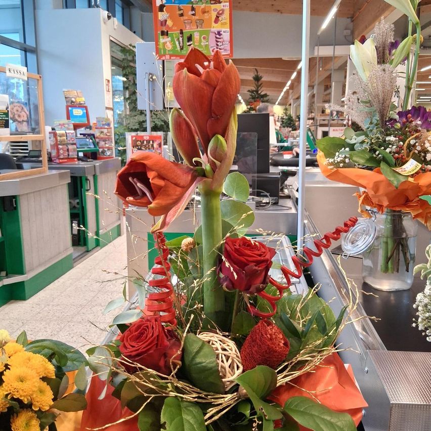 An orange flower arrangement with red roses and green leaves is on a checkout counter in a store. There are also other floral arrangements and products in the background.