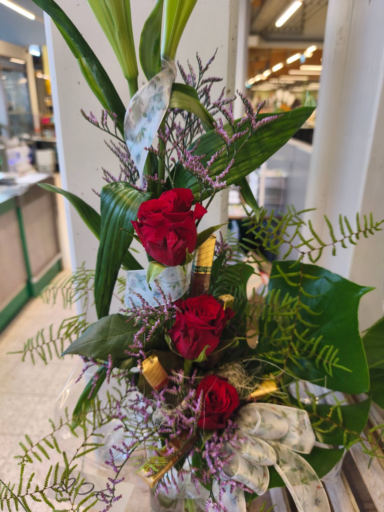 Eine Nahaufnahme eines Blumenarrangements mit drei roten Rosen, lila Blumen und verschiedenen grünen Blättern in einer kommerziellen Umgebung.