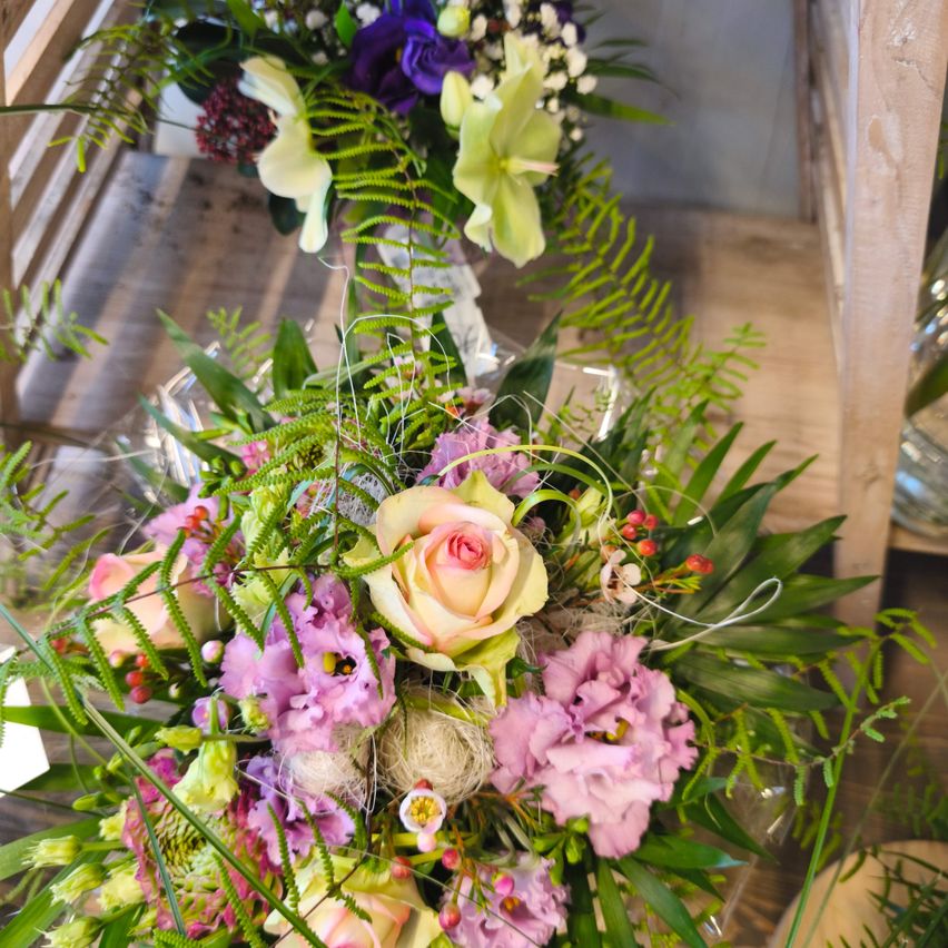 Eine farbenfrohe Blumenarrangement mit einer Mischung aus Rosen, lila und rosa Blumen und grünen Blättern, präsentiert auf einer Holzoberfläche.
