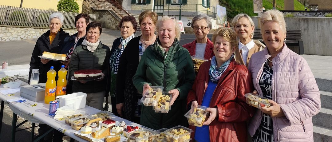 Eine Gruppe lächelnder Frauen steht vor einem gelben Gebäude mit einem Kirchturm. Sie halten Teller mit Essen und stehen neben einem Tisch mit Getränken. Der Himmel ist blau mit einigen Wolken.