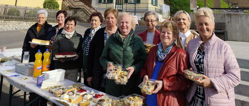Eine Gruppe lächelnder Frauen steht vor einem gelben Gebäude mit einem Kirchturm. Sie halten Teller mit Essen und stehen neben einem Tisch mit Getränken. Der Himmel ist blau mit einigen Wolken.