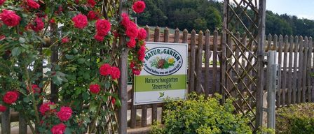 Ein Garten mit Rosen, einem Holzzaun und einem Schild, das Naturschaugarten Steiermark besagt. Das Gebiet ist von Bäumen umgeben.