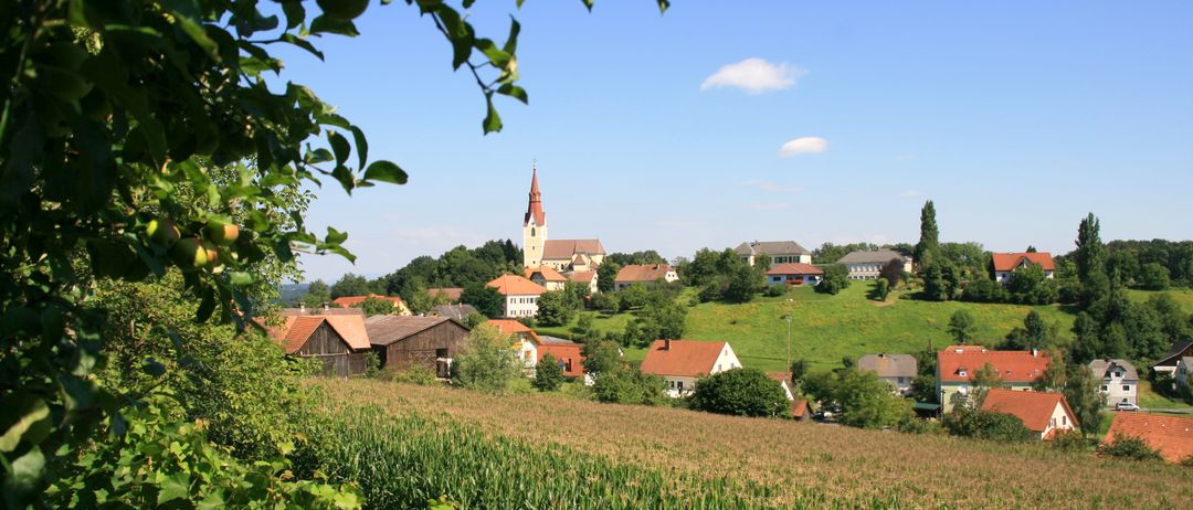 Ein ländliches Dorf mit Häusern und einer Kirche steht auf einem Hügel unter einem klaren blauen Himmel. Grüne Felder und Bäume umgeben das Dorf.