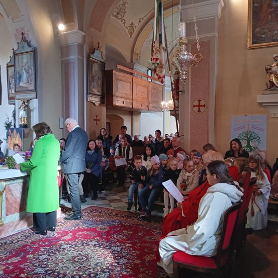 Eine Gruppe von Menschen, darunter eine Frau in einem grünen Mantel, versammelt sich in einer Kirche für eine Zeremonie. Die Kirche ist mit Gemälden, Fahnen und einer Kronleuchter dekoriert. Einige Menschen sitzen auf Stühlen, während andere stehen.