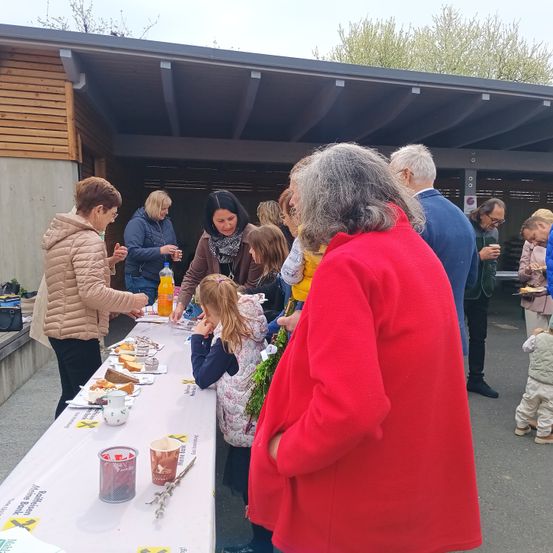 Mehrere Menschen stehen an einem langen Tisch mit Essen, Getränken und einem Becher mit Strohhalm. Eine Frau in einem roten Mantel steht vor dem Tisch, während andere hinter ihr stehen.