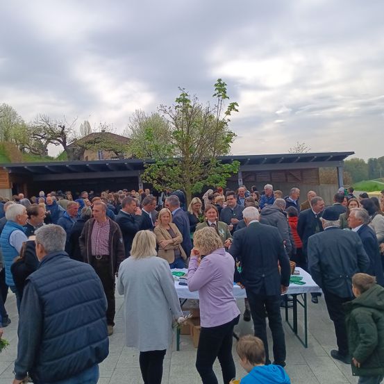 Eine Menschenmenge steht draußen, einige unterhalten sich, und in der Mitte der Gruppe befindet sich ein Baum mit einem langen Stamm.