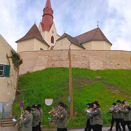 Eine Musikkapelle in Uniformen spielt Trompeten und Posaunen, während sie auf einer Straße vor einer Kirche marschiert.