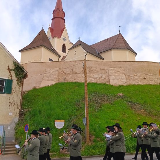 Eine Musikkapelle in Uniformen spielt Trompeten und Posaunen, während sie auf einer Straße vor einer Kirche marschiert.